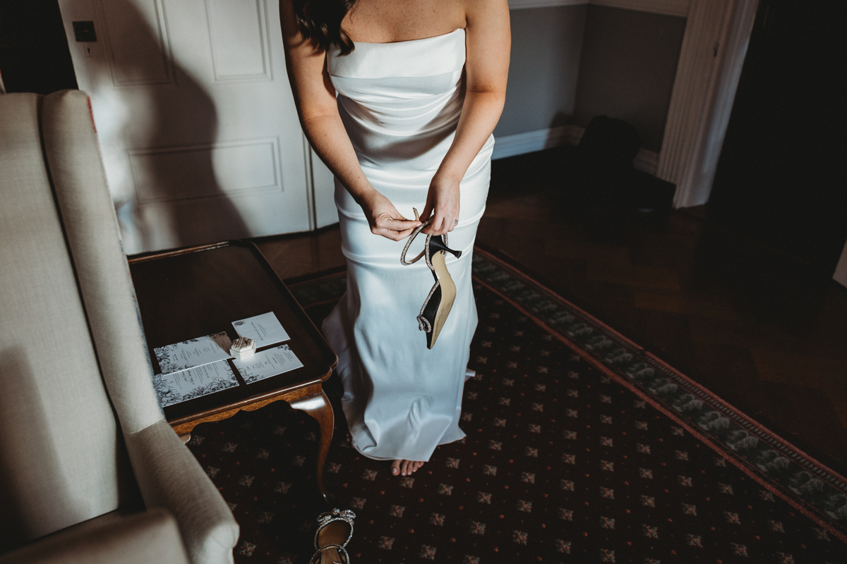 A bride adjusts her footwear while standing in her elegant wedding dress, with wedding invitations and a ring on a nearby table.