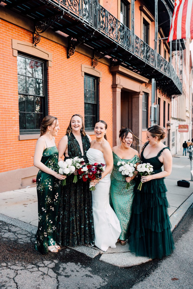 A group of five smiling bridesmaids in elegant dresses, each holding floral bouquets, standing together in front of a brick building and American flag.