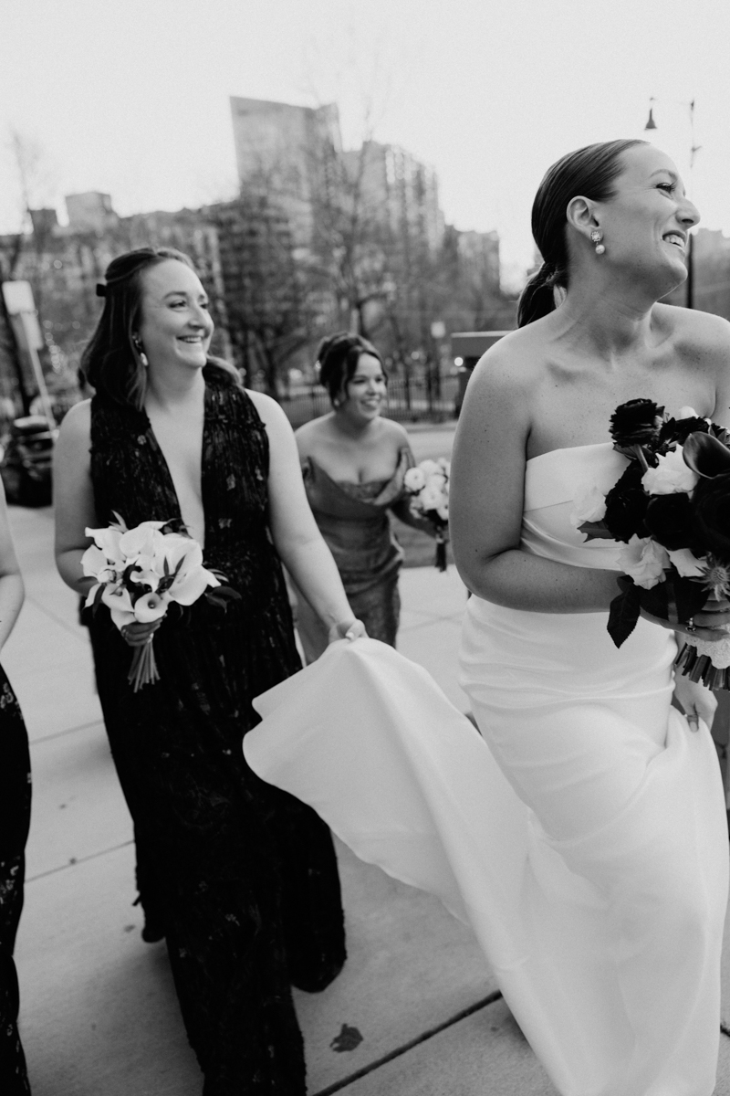 A joyful group of bridesmaids walking together in elegant dresses, carrying bouquets, and laughing in a city setting.