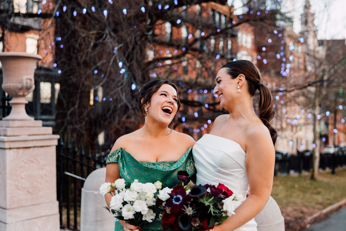 Two women, one in a green dress and the other in a white gown, share laughter while holding vibrant flower bouquets in a festive outdoor setting.