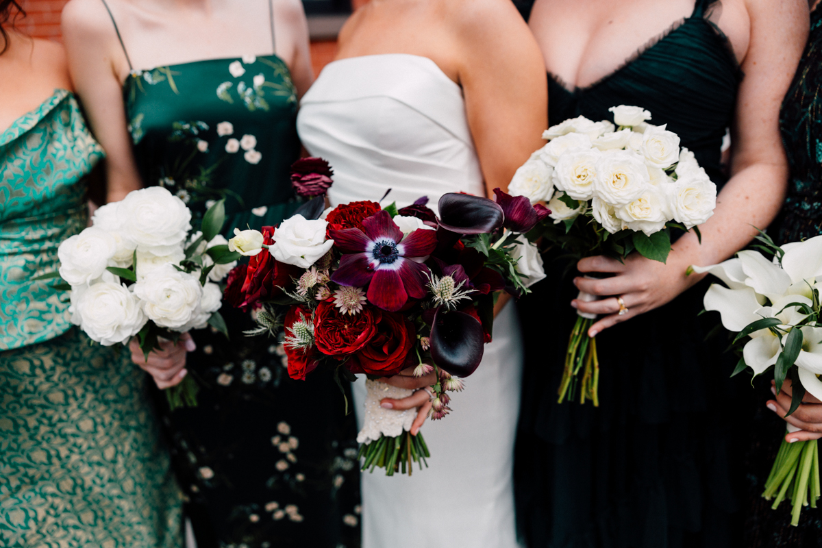 A group of women in elegant dresses holding various flower bouquets, showcasing a mix of blooms in rich colors, including white roses and deep reds.