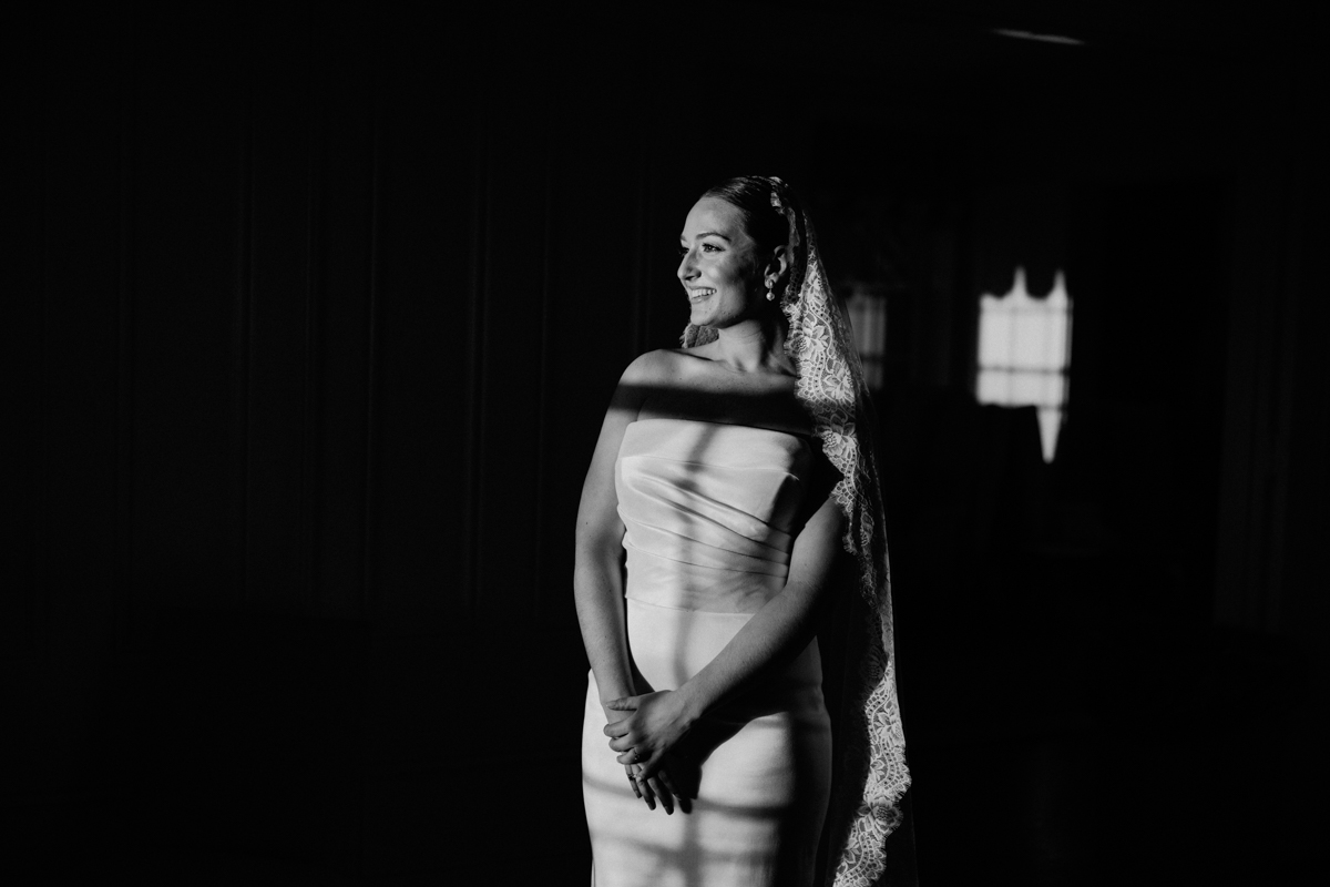 A bride dressed in a form-fitting gown and veil stands gracefully in soft light, casting delicate shadows on her figure.