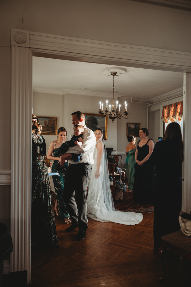 A man holding a baby amidst a group of elegantly dressed women in a well-lit room with vintage decor.