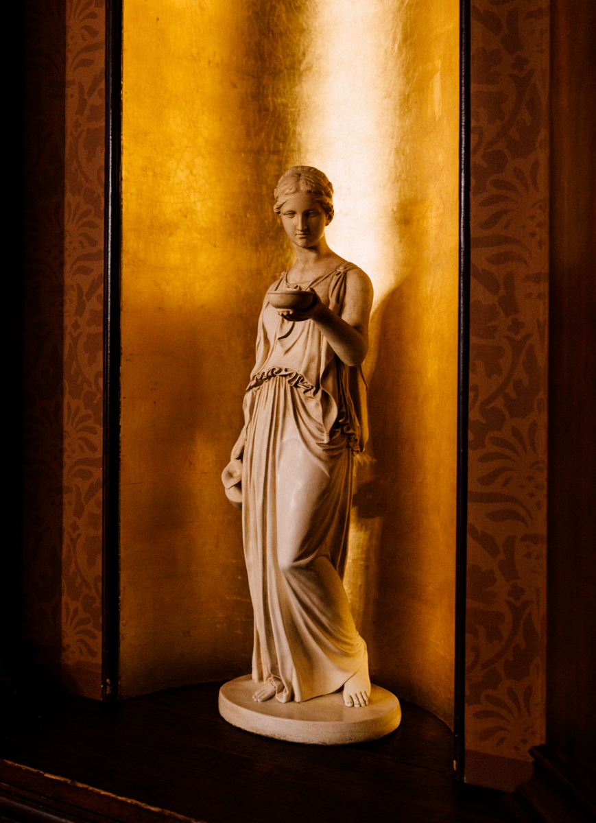 A graceful marble statue of a woman standing in a golden-lit niche, showcasing intricate details of her draped clothing and serene expression.