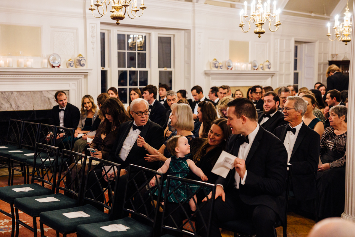 A gathering of elegantly dressed guests seated in a beautifully decorated room, engaging with each other, with a young child capturing attention in the foreground.