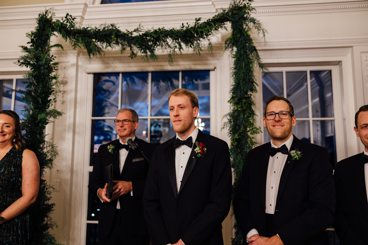 A formal gathering featuring a groom in a tuxedo and friends standing together by a decorated backdrop of greenery and large windows.