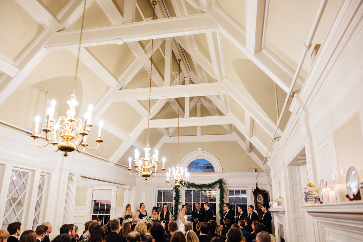 A bride and groom exchanging vows during a wedding ceremony in an ornate hall, featuring chandeliers and a decorated backdrop, surrounded by guests.