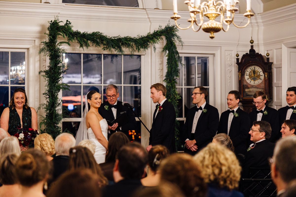 A bride and groom exchange vows during a winter wedding ceremony, surrounded by family and friends in a beautifully decorated room with greenery and a vintage clock.