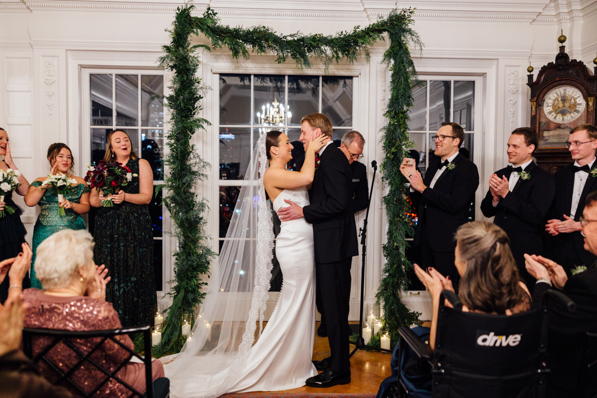 A joyful couple sharing a kiss during their wedding ceremony, surrounded by a decorated arch and applauding guests.