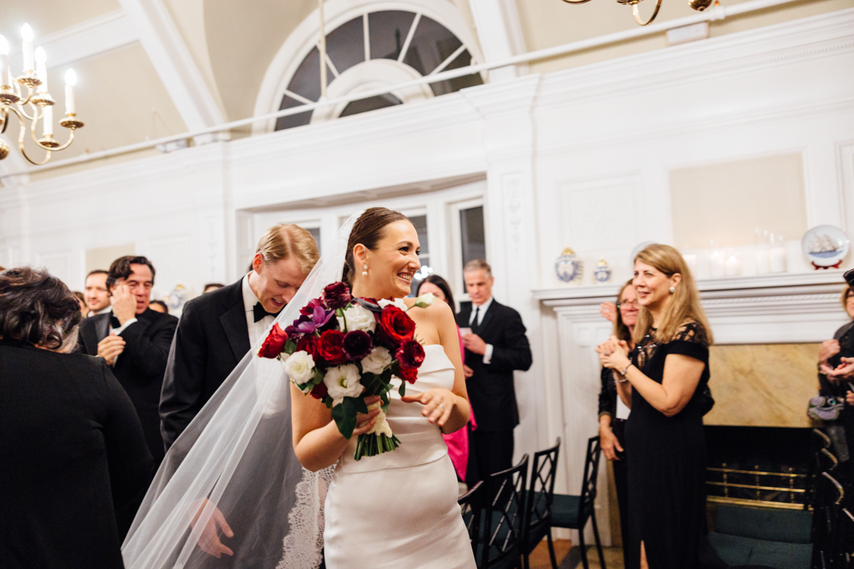 A bride smiles joyfully while holding a bouquet, as guests applaud in a beautifully decorated venue.