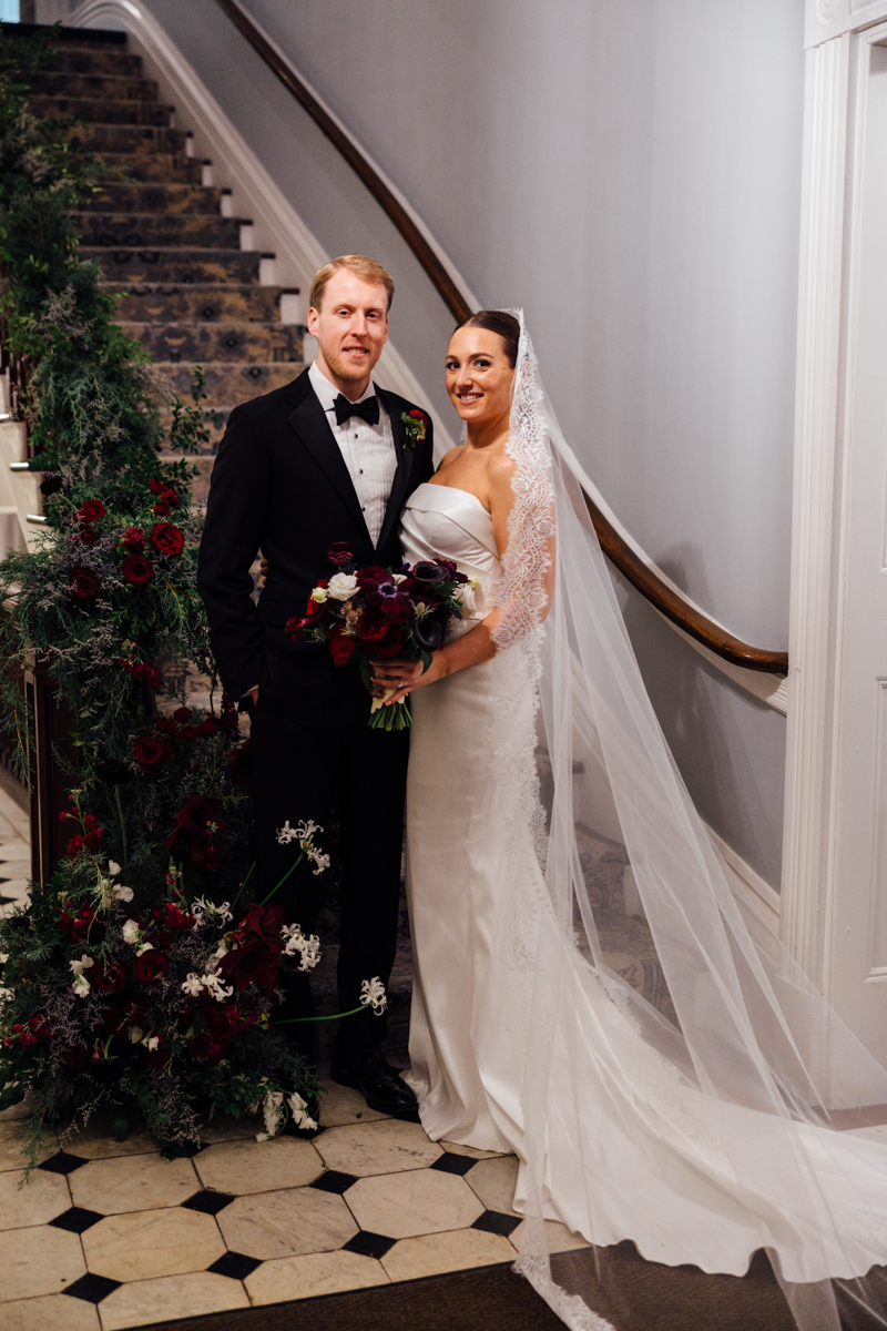 A smiling couple dressed in wedding attire stands together by a staircase adorned with floral arrangements, capturing a moment of joy and celebration.
