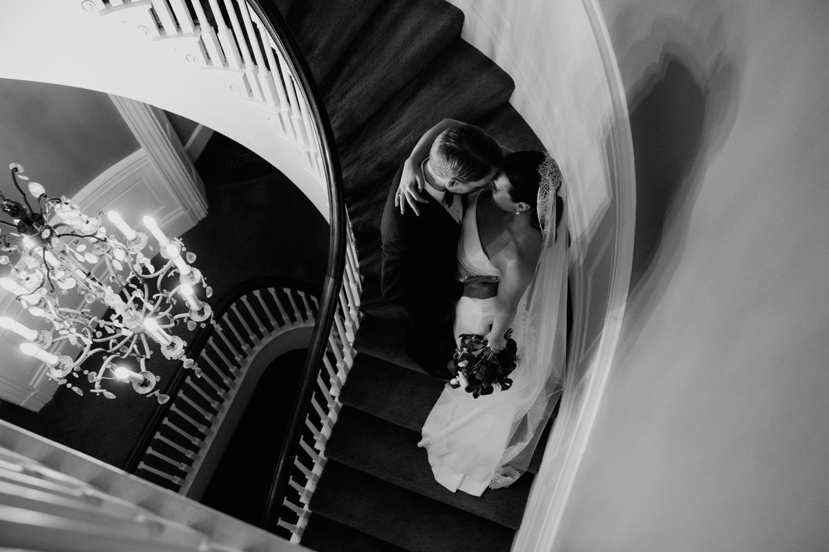 Bride and groom sharing a tender kiss on a spiraled staircase, illuminated by a chandelier, captured in black and white.