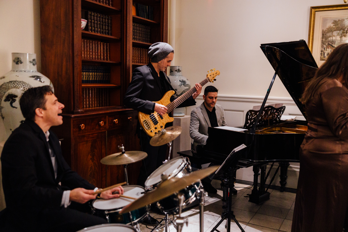 A jazz band playing in a sophisticated interior, featuring a drummer, bassist, and pianist amidst a backdrop of bookshelves and decorative vases.