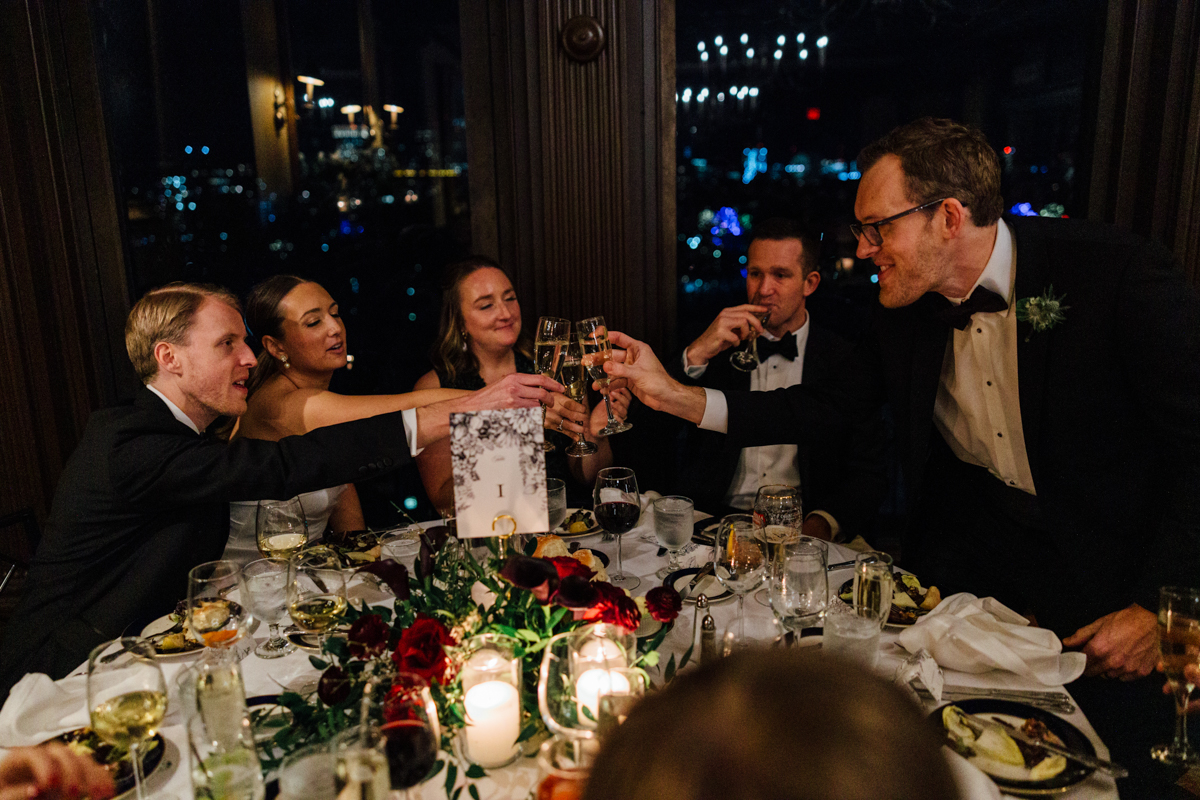 A group of elegantly dressed individuals clink champagne glasses over a beautifully set dinner table adorned with floral decorations and candles, overlooking a nighttime cityscape.