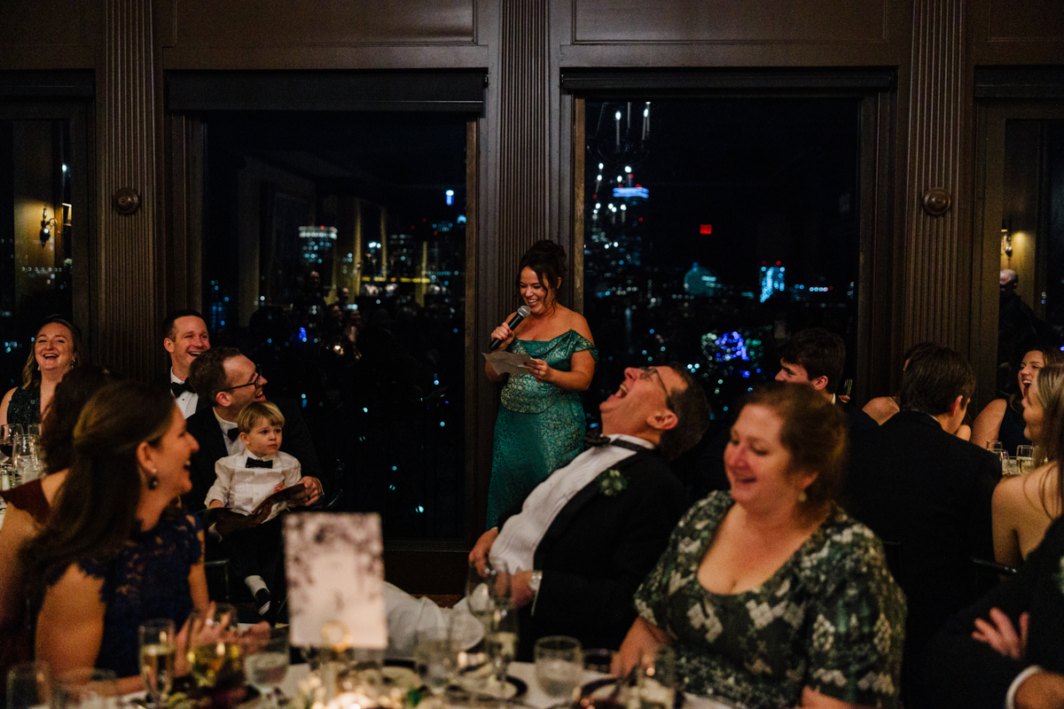 A woman in a green dress delivers a speech while guests enjoy the evening, with city lights shining in the background.