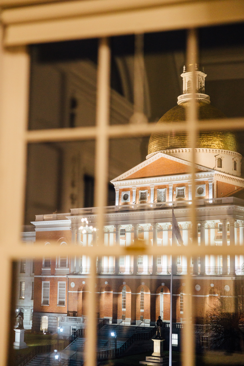View of the illuminated Boston Statehouse through a window at night, with a focus on its golden dome and architectural details.