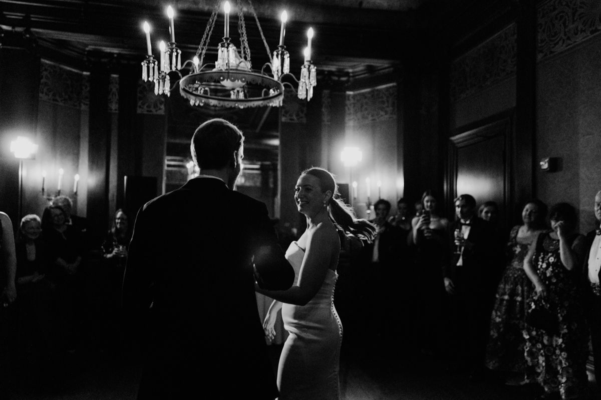 A couple dances closely in a dimly lit room, surrounded by their guests, conveying an atmosphere of joy and celebration.
