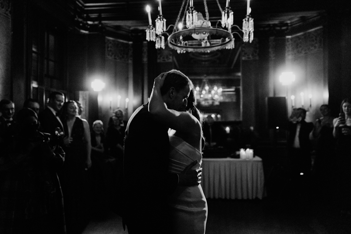 A couple shares a romantic dance in a dimly lit hall, surrounded by guests capturing the moment.