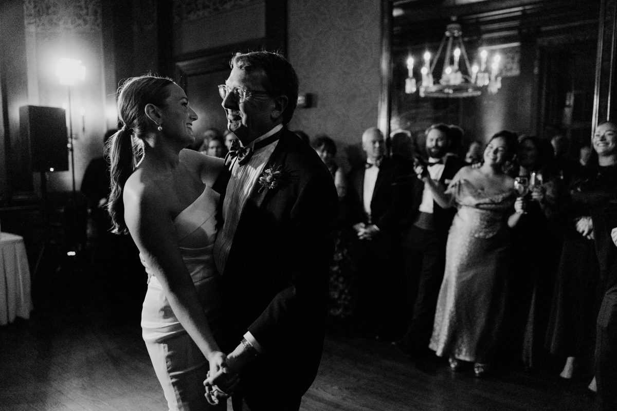 A joyous father-daughter moment captured in black and white during a wedding reception, with guests in formal attire watching in the background.