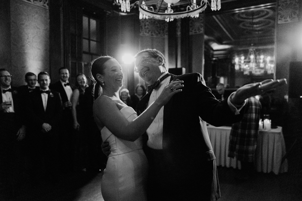 A groom dances with his daughter, both smiling widely, surrounded by guests in formal attire, capturing a moment of joy and celebration in a beautifully lit venue."