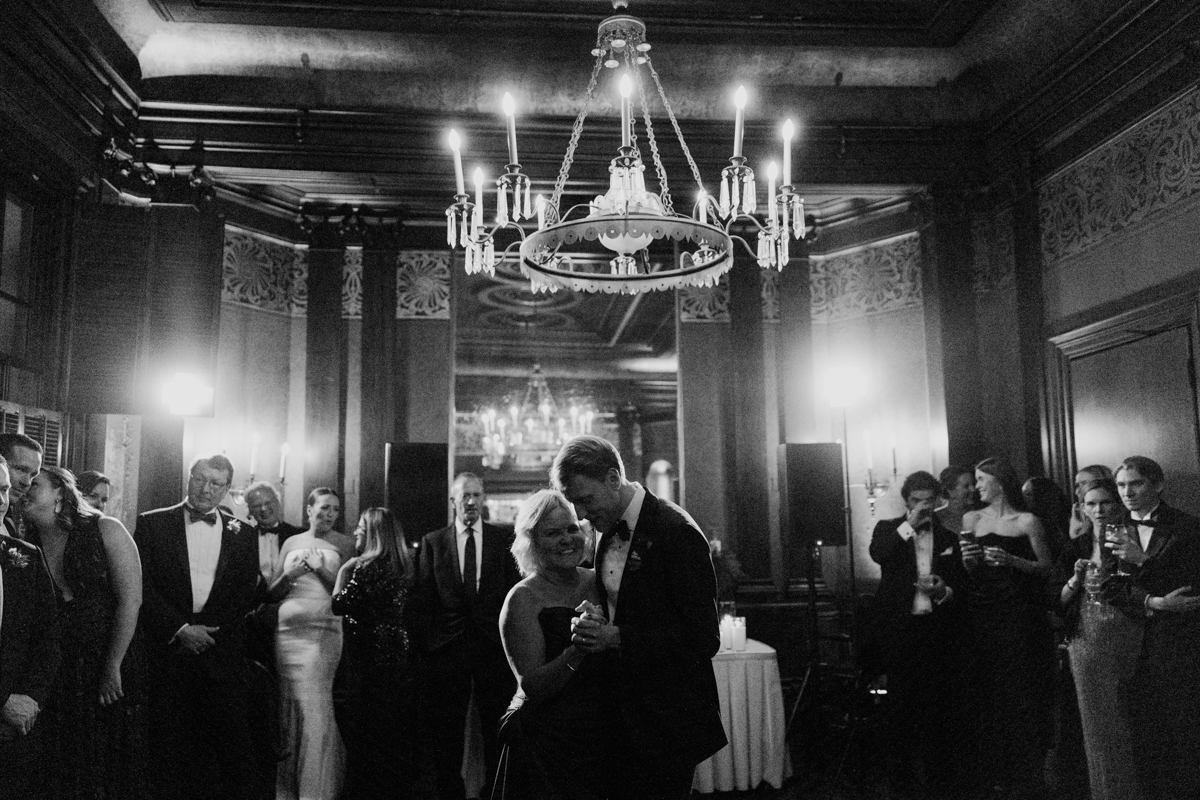 A couple shares a heartfelt moment on the dance floor during a wedding reception, surrounded by elegantly dressed guests in a beautifully decorated venue.