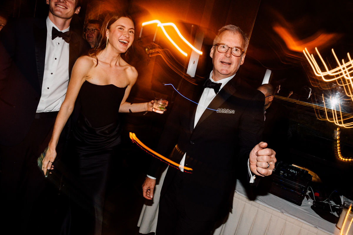 A lively scene of three elegantly dressed individuals at a festive event, with one woman laughing joyfully while holding a drink, and a man in a tuxedo gesturing, surrounded by vibrant light trails.