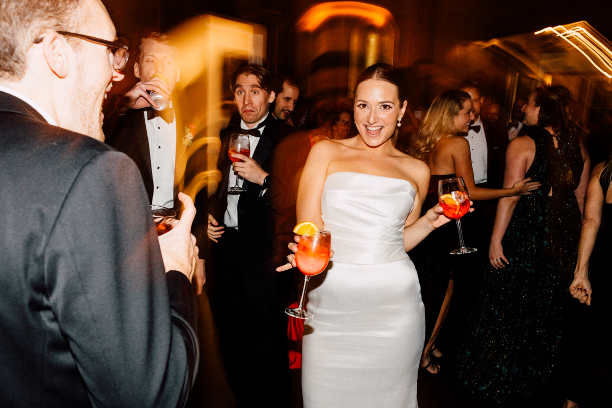 A lively party scene featuring a woman in a white gown joyfully holding a colorful drink, surrounded by elegantly dressed guests enjoying the festivities.