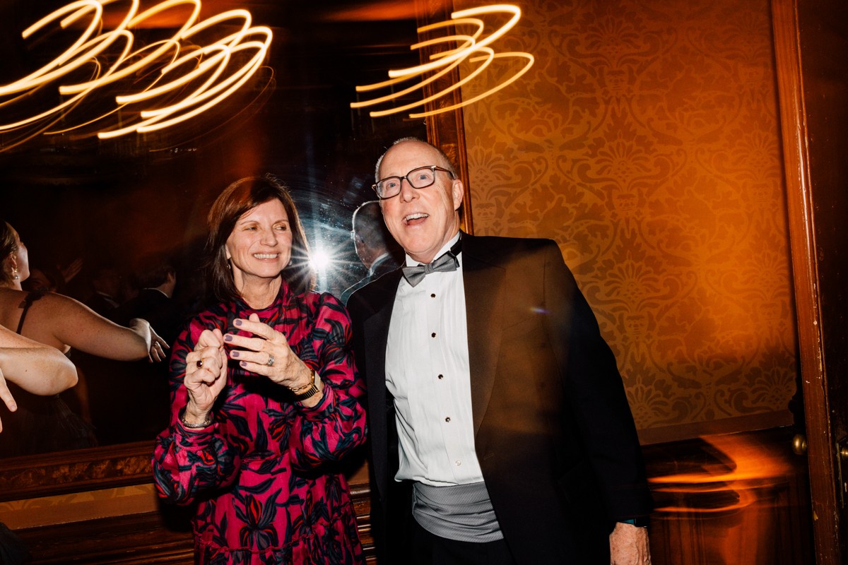 A man in a tuxedo and a woman in a vibrant floral dress share a joyful moment, surrounded by blurred lights and elegant decor in a festive atmosphere.