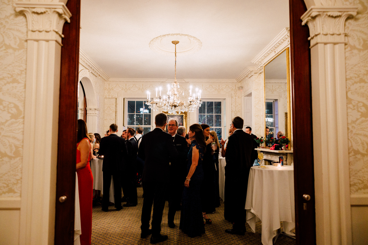 A stylish indoor setting featuring a group of guests mingling at an event, illuminated by a chandelier, with decorative elements and a warm atmosphere.