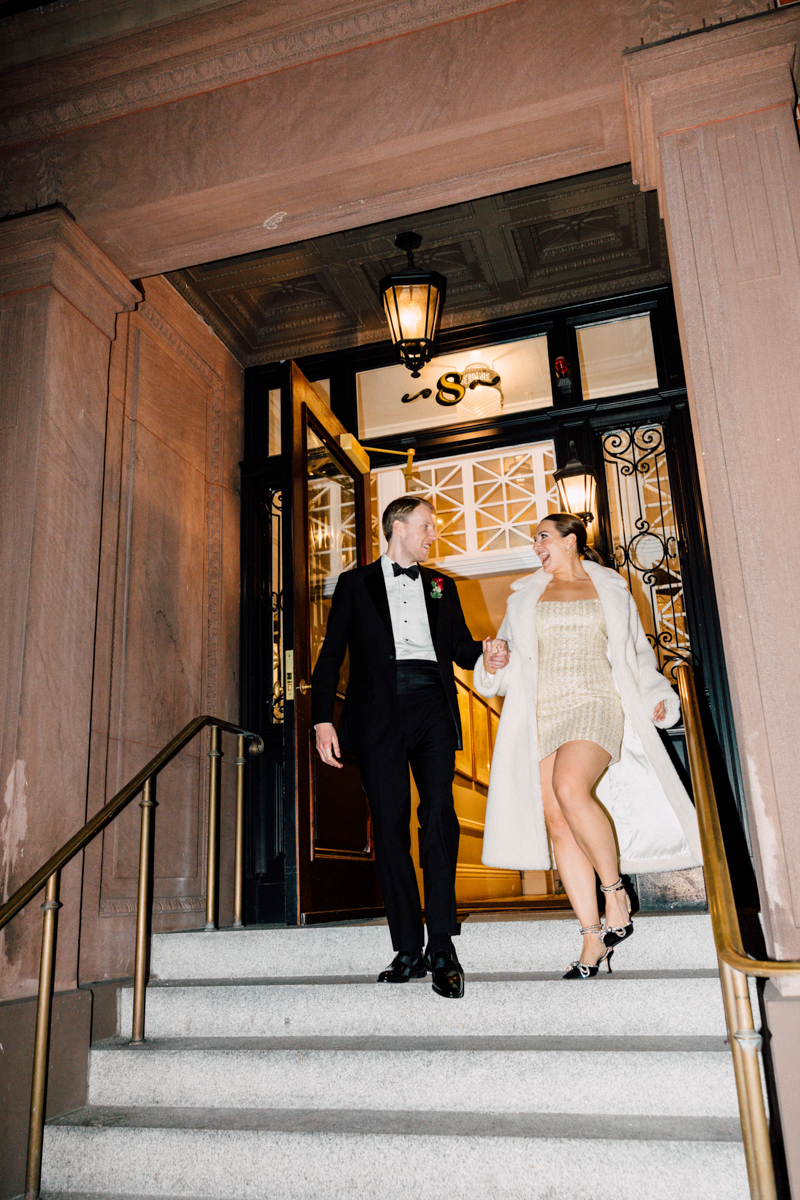 A stylish couple descends the stairs of a grand venue, dressed elegantly for a night out, with the woman wearing a shimmering dress and a warm coat.