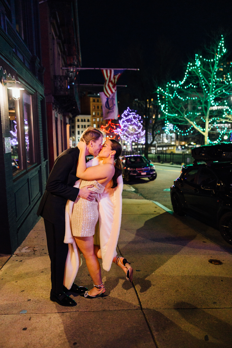 A couple sharing a passionate kiss on a city street adorned with colorful holiday lights, surrounded by a vibrant night atmosphere.