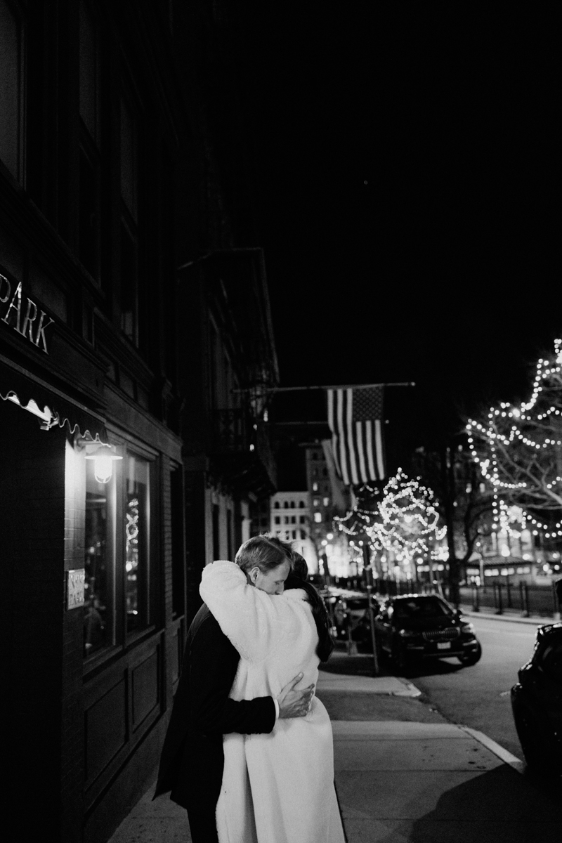 A couple shares a warm embrace on a street adorned with festive lights, illuminated by a gentle glow from a nearby building.