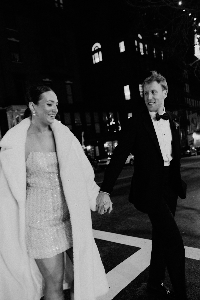 A happy couple walks hand in hand on a city street at night, with the woman in a sparkling dress and fur coat, while the man wears a tuxedo.