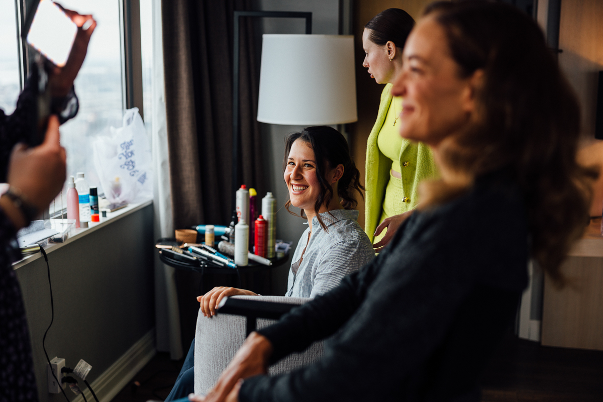 A woman smiling in a chair while getting ready, with friends assisting her in a bright, stylish room filled with beauty products.