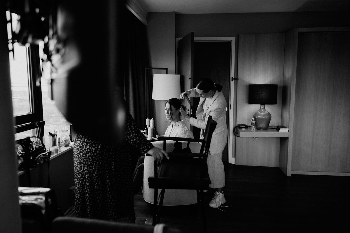 A woman smiles as a stylist works on her hair in a stylish hotel room, with natural light illuminating the scene.