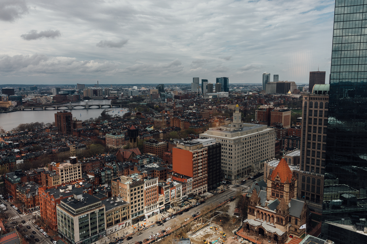 Aerial view of Boston's downtown skyline with a mix of historic and modern architecture, showcasing the Charles River in the background under a cloudy sky.