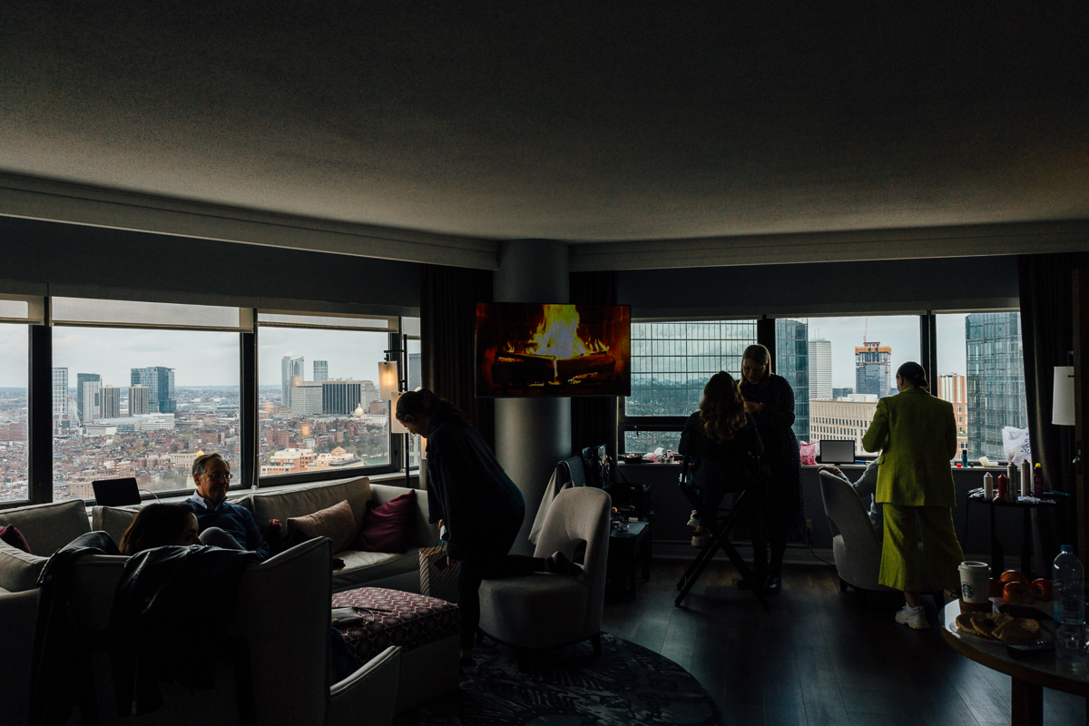 A group of people preparing makeup in a stylish hotel room overlooking a bustling cityscape with a cozy fireplace display on the TV.