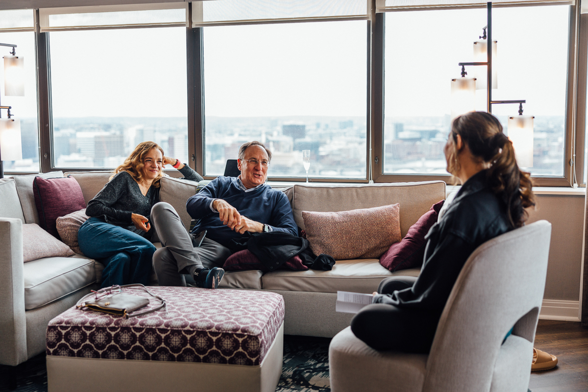 A man and woman engage in a light-hearted conversation on a couch while another woman listens attentively in a stylish lounge with a city view.