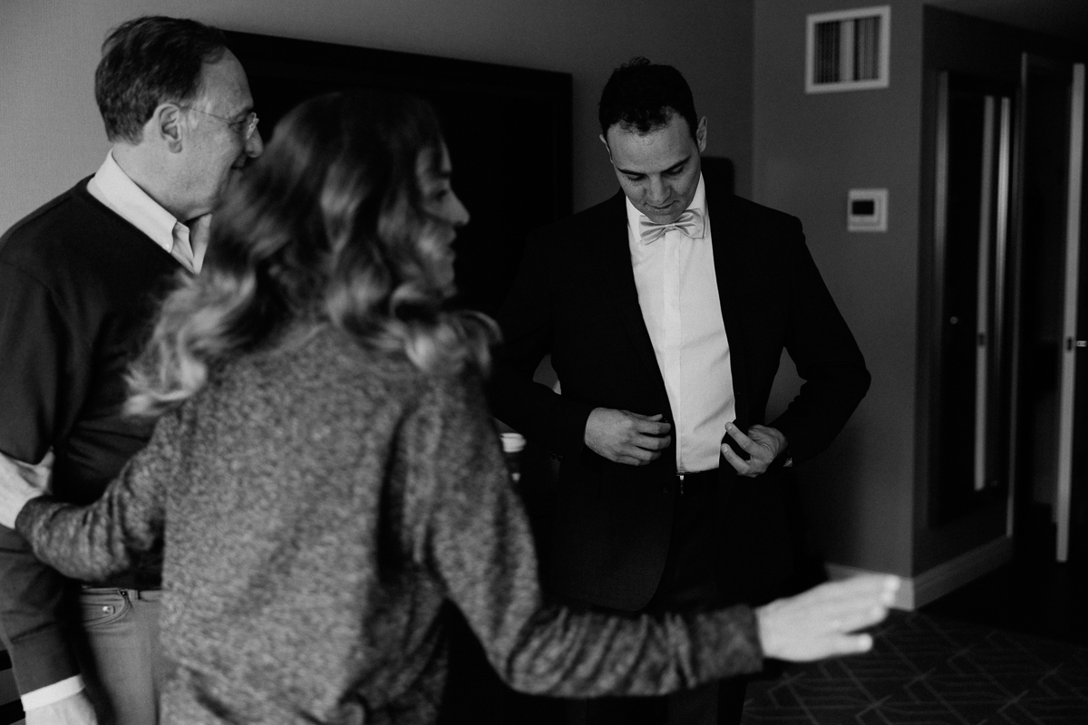 A groom in formal attire adjusts his bow tie, while a woman in a sweater and a man in casual clothing engage in conversation, capturing a moment of pre-wedding excitement.