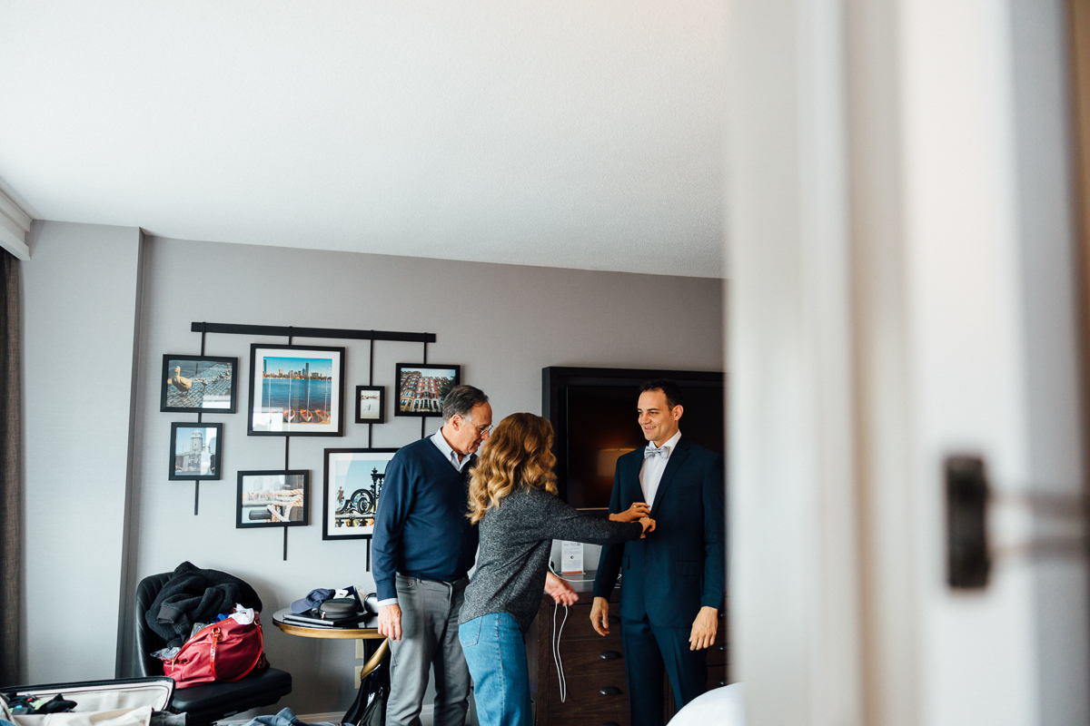 A groom receives help adjusting his attire in a hotel room while two assistants offer support and encouragement.