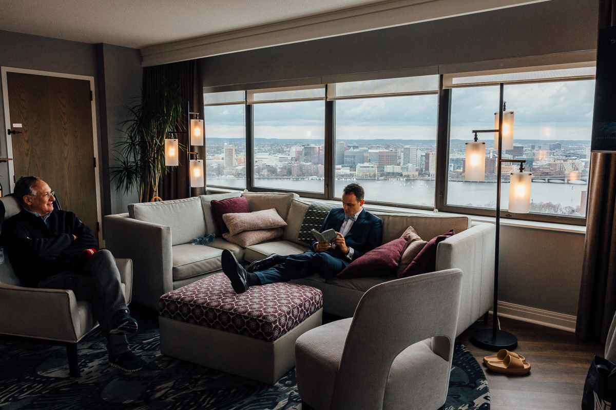 A man reading in a modern living room with a view of the city, while another man sits nearby, both in a stylish, comfortable setting.
