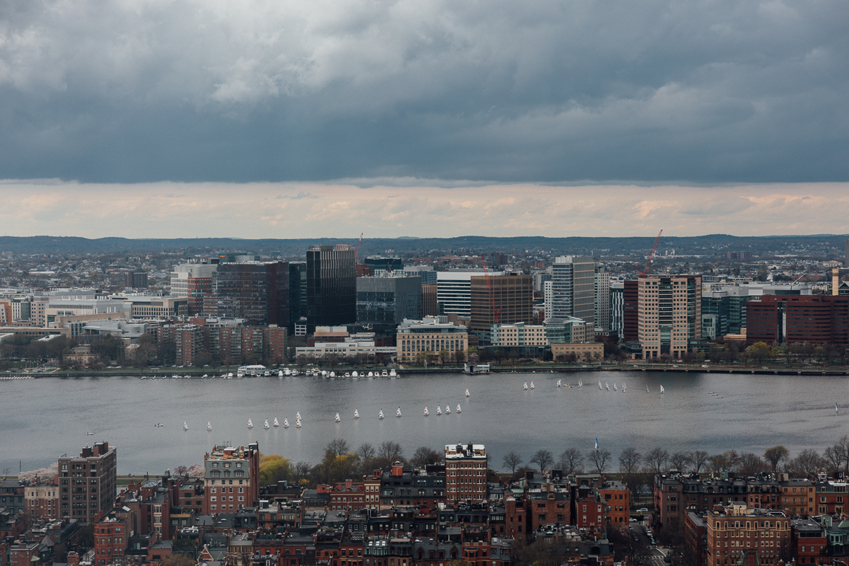 Aerial view of Boston's skyline with sailing boats on the Charles River under a cloudy sky.
