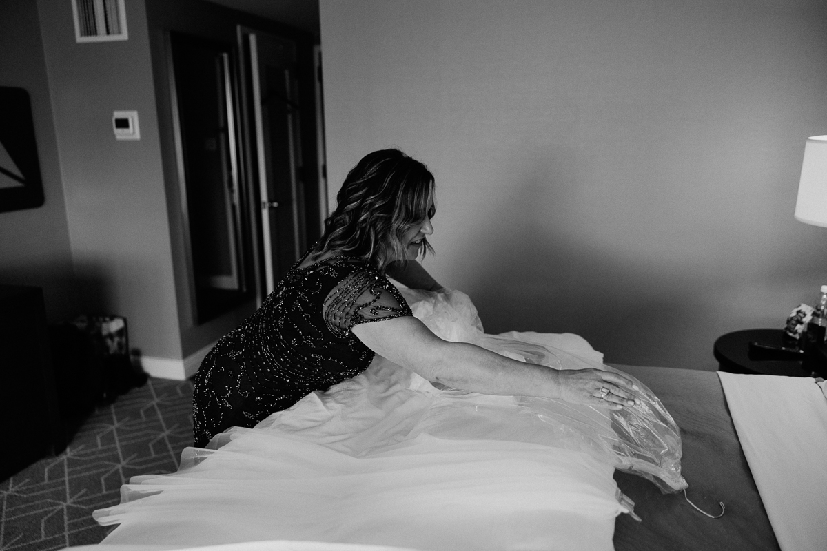 A woman in a beaded black dress carefully arranges a wedding gown on a bed in a hotel room, focusing on the delicate fabric.