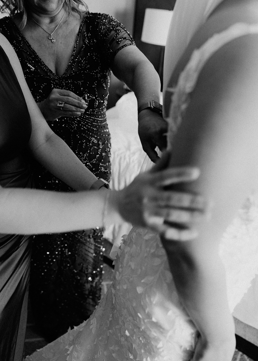A close-up of hands assisting a bride in her wedding gown, showcasing moments of support and intimacy before the ceremony.