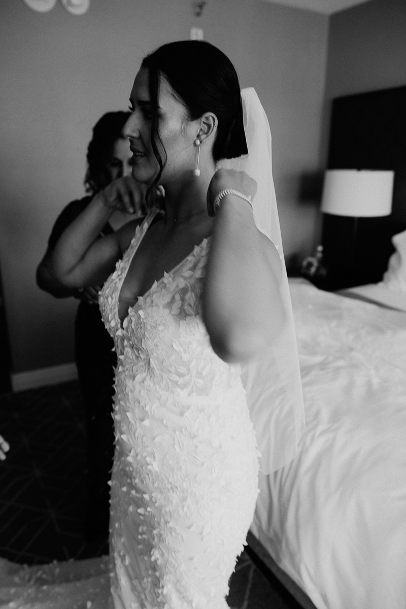 A bride adjusts her veil while dressed in a floral wedding gown, with a friend assisting her in a hotel room.