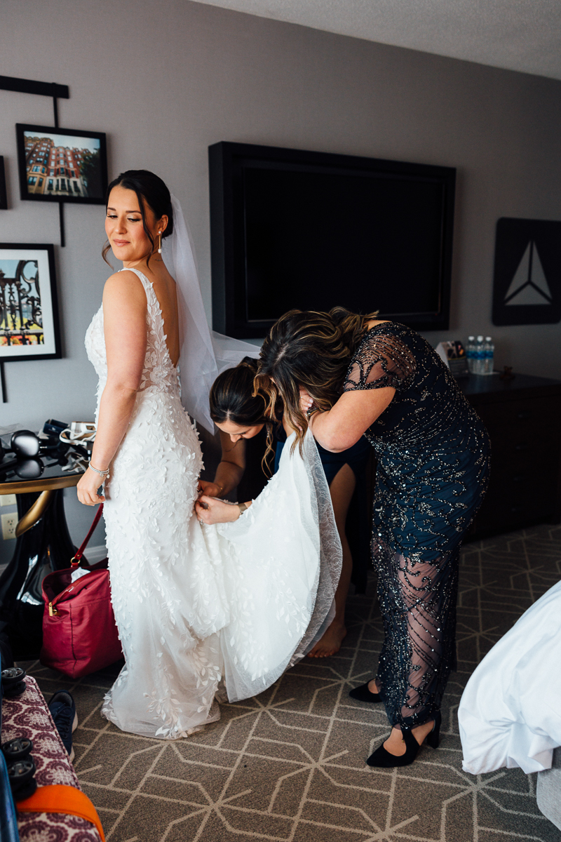A bride smiles as her friends assist her in adjusting her wedding dress in a hotel room, filled with excitement and anticipation.