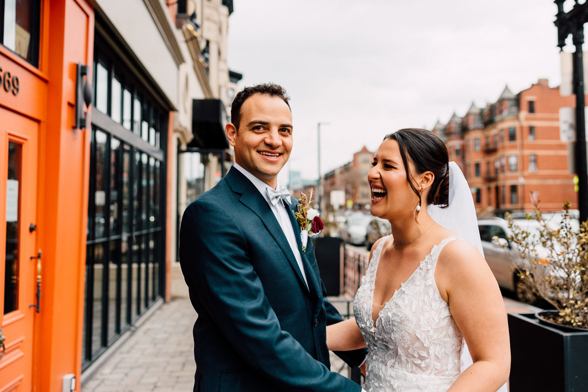 A happy couple smiles and laughs together outside in a lively city street, celebrating their wedding day.