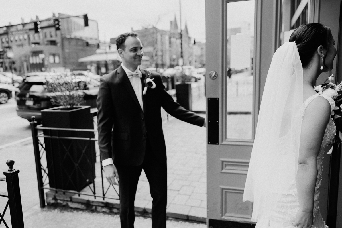 A groom smiles as he opens the door for his bride, moments before their wedding ceremony, capturing a joyful and intimate moment on a bustling street.