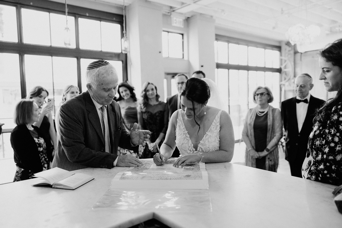 Bride signing a ceremonial document with witnesses in an elegantly decorated venue.