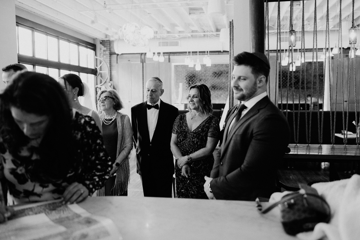 A group of well-dressed individuals capture a moment at a stylish venue, with expressions of anticipation and joy as one person focuses on writing at a table.